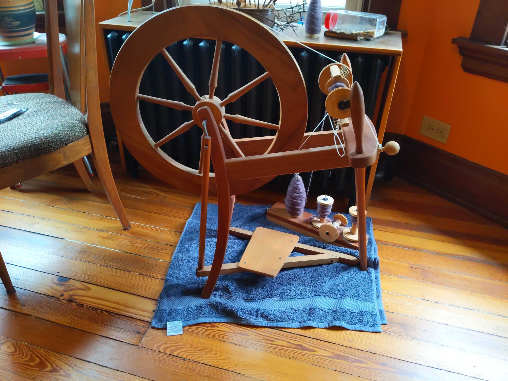 A spinning wheel sitting on a blue towel in the middle of a wood floor with a purple fiber being spun.