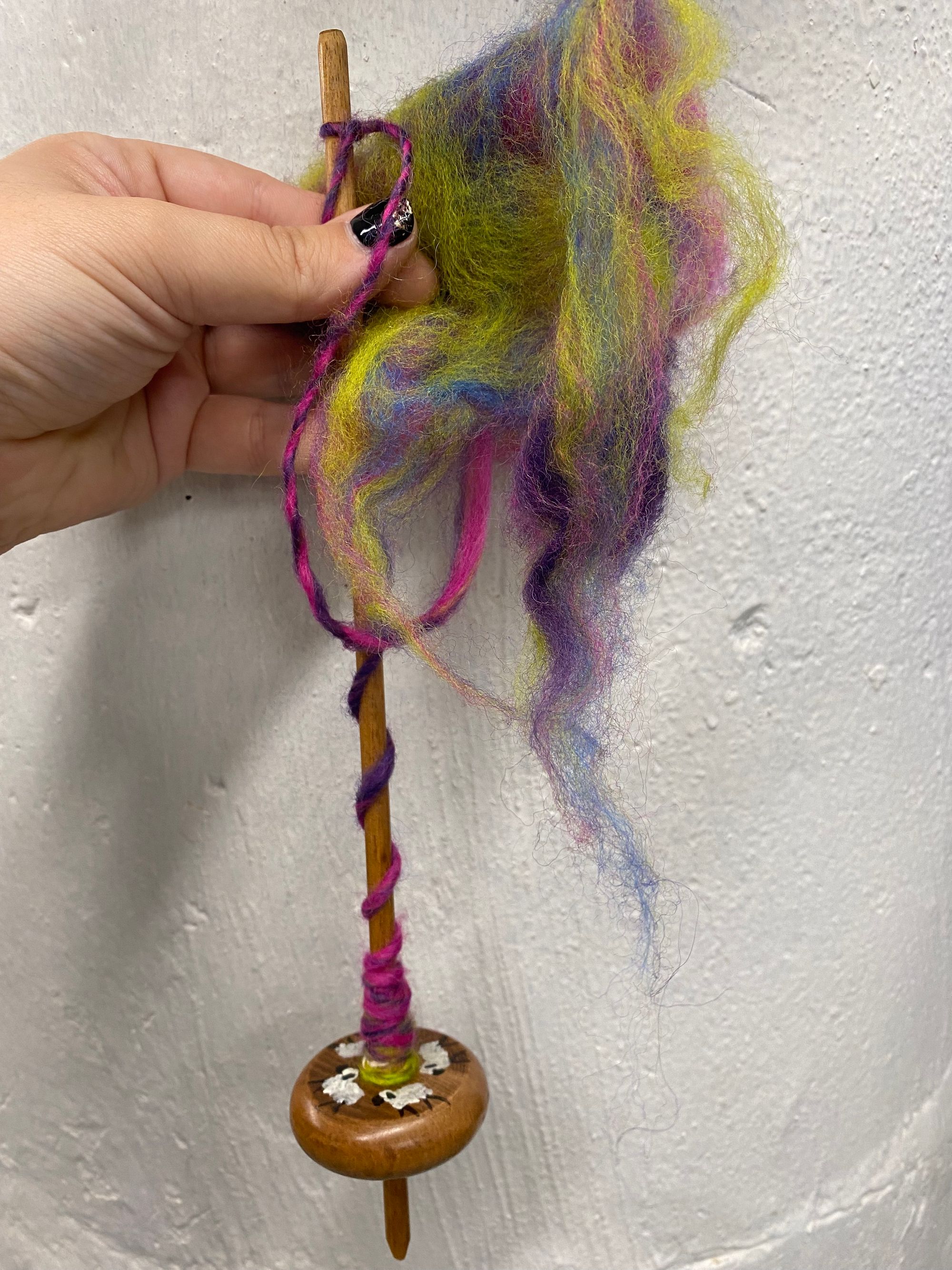 A drop spindle with little sheep painted on it held against a white, concrete surface with green, purple, and pink fibers coming off of it.